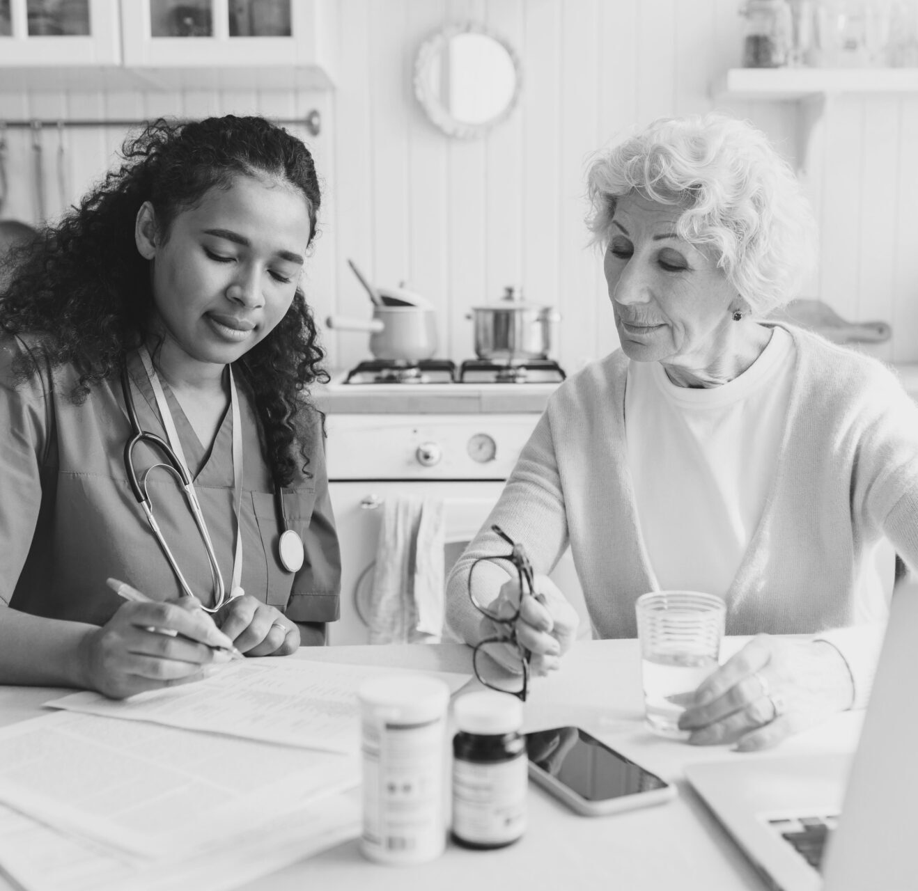 Elderly lady sitting at kitchen table near african american nurse in blue uniform writing prescriptions after giving her first aid, senior woman looking attentively at notes, holding bottle of water